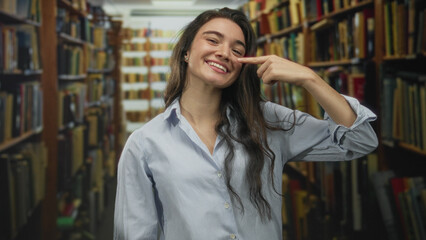 Young hispanic woman pointing finger to nose among tall bookshelves in old library building indoors  thoughtfulness. © Krakenimages.com