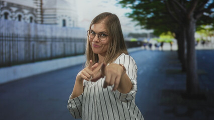 Woman pointing finger to camera in urban street with blurred building facade and tree lined walkway; confidence.