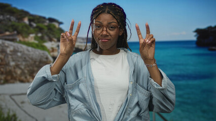 Woman wearing glasses and denim shirt holding hands in air quotes gesture at studio seaside set with turquoise ocean backdrop; skepticism irony.