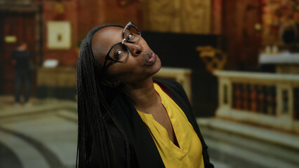 Fototapeta premium African american woman with glasses smiling joyfully indoors at a church setting with elegant decor in the background.
