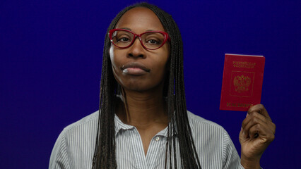 Woman holding russian passport in front of blue background, wearing glasses and striped shirt,...