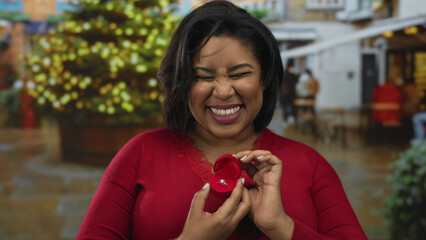 Woman smiling holding red ring box outdoors with festive decorations, capturing the essence of joy, latin culture, and a heartfelt proposal moment in a vibrant city setting.
