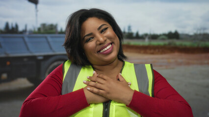 Woman in reflective vest smiling with hands on chest outdoors on city street, showcasing positivity and confidence.