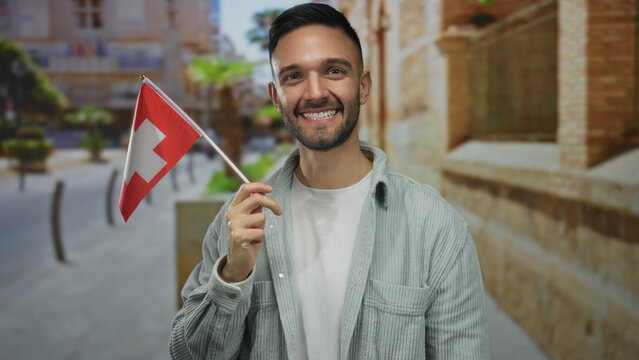 Young man smiling and holding a switzerland flag standing outdoors in an urban city street setting showcasing multicultural pride and diversity in a vibrant cityscape.