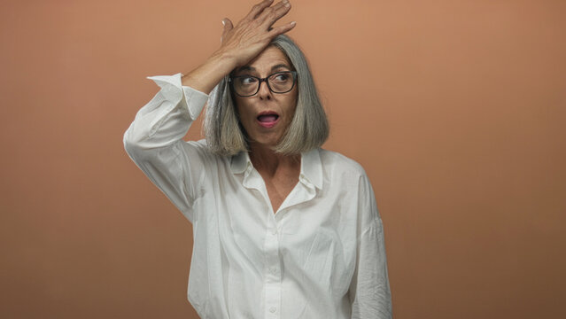 Woman with gray hair holds hand to forehead in studio with peach backdrop and white shirt; surprise realization.