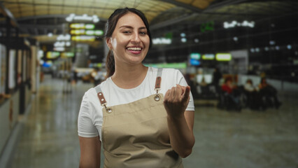 Woman wearing beige apron beckoning with hand in busy airport terminal near seating area and...