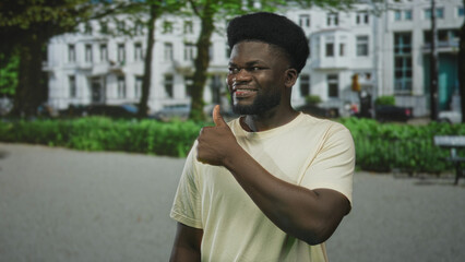 Fototapeta premium Man showing thumbs up on street in front of white apartment building and green park hedges, smiling with bare hand and thumb visible; confidence positivity.