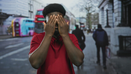 Fototapeta premium Man covering eyes and cupping ears with hands on a busy city street beside a red doubledecker bus wearing a red polo; playful curiosity.