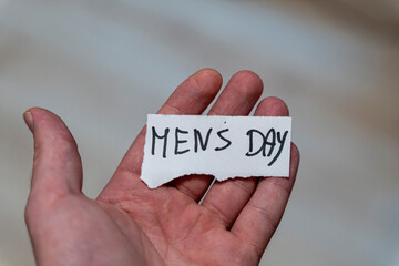 A hand holding a paper with handwritten words Men's Day. The background is blurred, emphasizing the importance of recognizing men and their roles in society. Applicable for various celebrations.