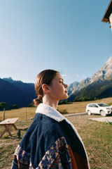 Fototapeta premium Woman profile in mountains field by a roadside car, wearing a warm jacket and feeling sunlight on her face, outdoor portrait of a young traveler standing near picnic table under clear sky