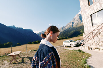 Fototapeta premium Woman standing near a stone chalet by a parked car in a grassy meadow, serene mountains in background. Outdoor portrait, travel lifestyle and rural countryside scene.