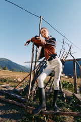 Naklejka premium Woman model in countryside wearing sweater and rubber boots, striking a confident pose while leaning on rustic fence with mountains and open field in natural sunlight.