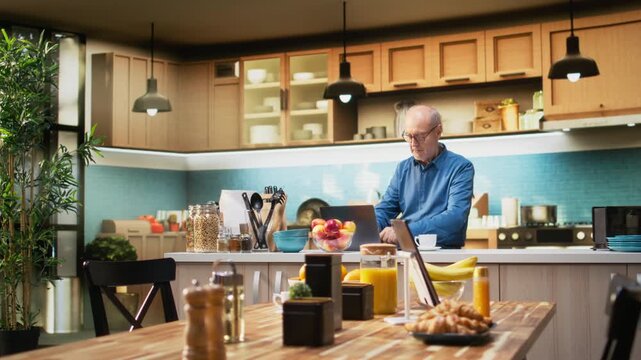 Senior adult in calm kitchen setting scrolling apps on her laptop, enjoying peaceful retired life. Old man showing gentle smile, simple moments that reflect relaxed domestic living. Camera A.