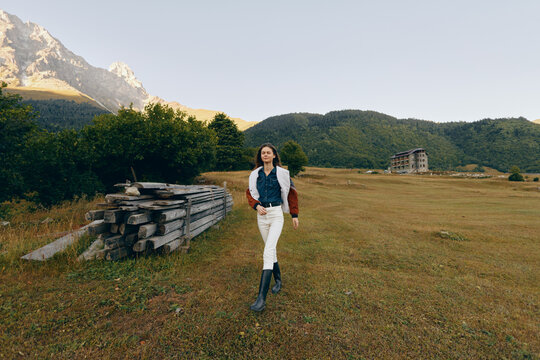 Woman walking in a grassy field near mountains and a wood pile, countryside scene with jacket and boots, autumn light and scenic nature for travel and outdoor lifestyle.