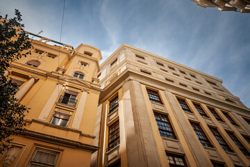 Low-angle view of residential facades in Valencia Ciutat Vella in spain. Classical details, tall windows, and light yellow tones characterize the old town streetscape amid bright sky and urban density © Jerome