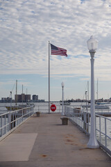 American Flag Flutters Over Calm Pier By Sea With Boats And White Lamps in Gulfport, FL. Serene waterfront scene featuring an American flag on a tall pole, white decorative lamps, a wide pier, boats i