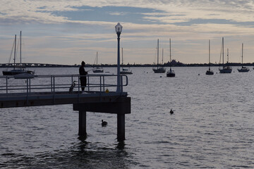 Harbor Pier At Dusk With Silhouette Fisherman, Sailboats And Calm Water Scene in Gulfport, FL. Distant sailboats. Gentle mood with subtle light and reflective surface.
