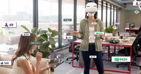 Seated woman tapping tablet, colleague donning VR headset causing UI badges rising in tech demo