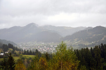 Misty Verkhovyna village nestled among rolling mountains and dense pine forests. Overcast skies loom above, creating a tranquil rural landscape with distant houses and greenery. Carpathians