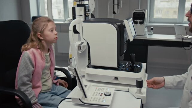 Side view shot of male doctor as optometrist changing configuration of automated diagnostic machine while performing eye exam for young girl in clinic, focus flowing from man to patient