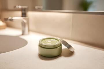 Green cream container sits on a bathroom counter near a sink and faucet in a well-lit room
