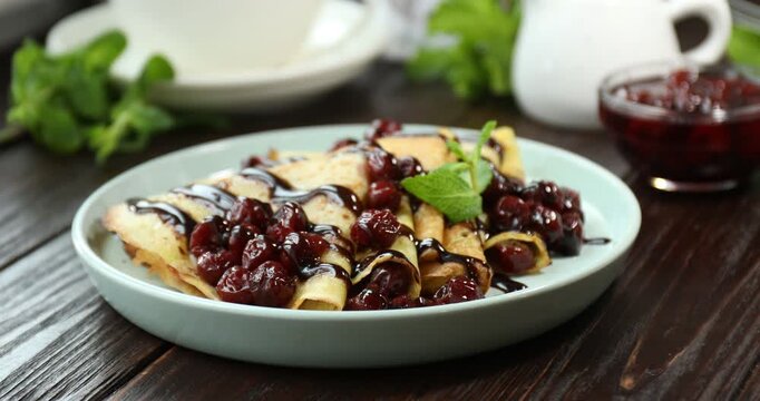 Woman adding mint to tasty crepes with cherries and chocolate at wooden table, closeup