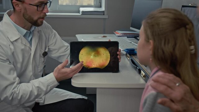 Over shoulder shot of male doctor as ophthalmologist pointing at images on digital tablet screen while consulting family with child in eye clinic