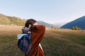 Fototapeta premium Woman in patterned sweater stretches and relaxes in nature on a grassy meadow with mountain landscape in the background, outdoors travel portrait capturing calm and freedom.