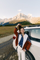 Obraz premium woman car mountains travel smile road nature adventure portrait of a happy female by an SUV, wearing casual jacket and denim, standing on a scenic roadside enjoying outdoor freedom.