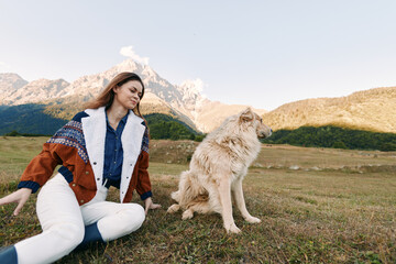 Fototapeta premium Woman and dog sitting in a mountain meadow outdoors, casual autumn jacket, enjoying nature and pet companionship, relaxed portrait with scenic peaks and open grassy field.
