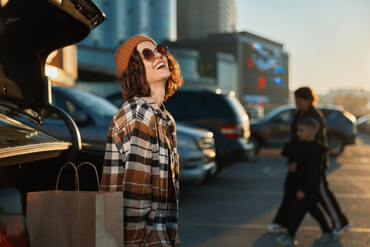 Woman shopping smile sunglasses coat car parking urban scene by trunk with shoppingbag, beanie and plaid, golden hour glow, authenticity, candid lifestyle, mindful living and emotional storytelling