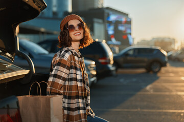 Obraz premium Woman shopping at car in parking lot with bags, smile and sunlight; candid lifestyle portrait with authenticity, mindful living and golden hour glow capturing emotional storytelling.