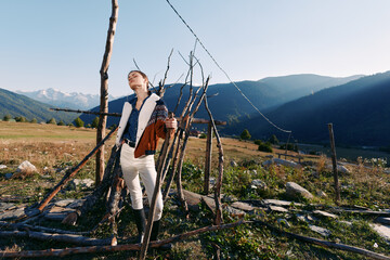 Fototapeta premium Woman in countryside near rustic fence and meadow, portrait enjoying sunlight with mountains in background and wide open field. Outdoor rural lifestyle, fashion and peaceful nature scene.
