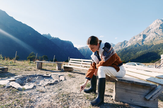 Woman sitting on a wooden bench in autumn mountains, wearing rubber boots and warm jacket in an outdoor lakeside campsite, touching ground and enjoying peaceful nature with clear sky. - Powered by Adobe