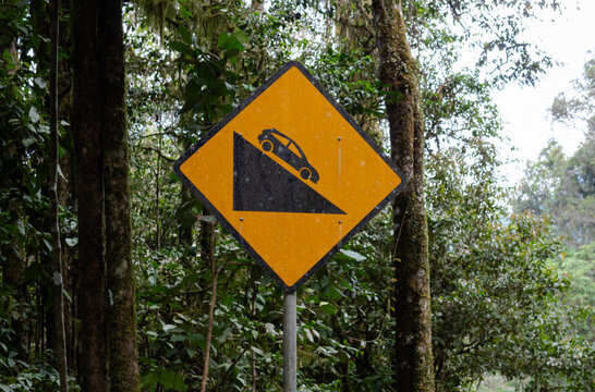 Bright yellow warning sign indicating steep downhill slope caution for drivers in forest