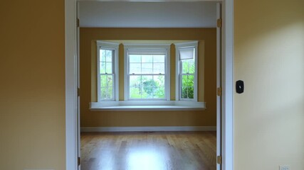 Empty living room in a residential house featuring warm yellow walls, polished hardwood floors, and large windows that fill the space with soft natural light.