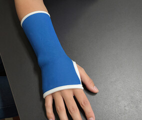 A person's arm and hand rest on a dark gray surface while wearing a fitted blue compression sleeve or forearm support with white trim.