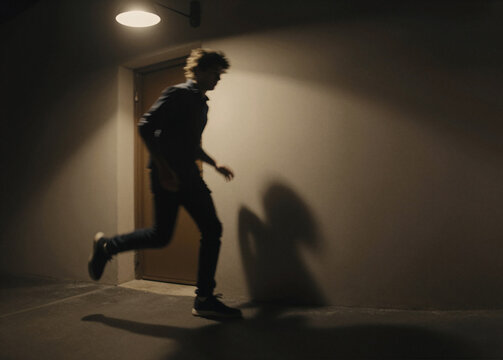 A silhouetted young man runs in a dimly lit corridor, creating a large, dark shadow on the adjacent wall.