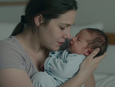 A light-skinned adult woman gently holds a sleeping newborn baby close to her face, showing a bond of maternal love in a soft, quiet setting.