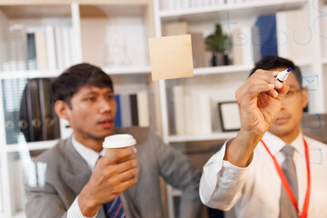 Two male employees in suits are designing a business plan, discussing various plans in their office, and one is drawing on a whiteboard. The scene looks cheerful and fun.

