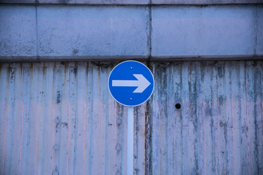 A blue circular traffic sign with a white arrow mounted on a pole in front of a ridged concrete wall.