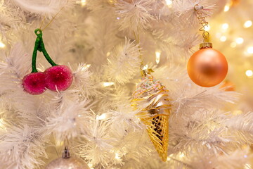 Cute pastel Christmas ornaments, including a pink cherry, golden ice cream cone, and peach bauble, hanging on a white tree with warm glowing lights. A whimsical and festive holiday close-up.