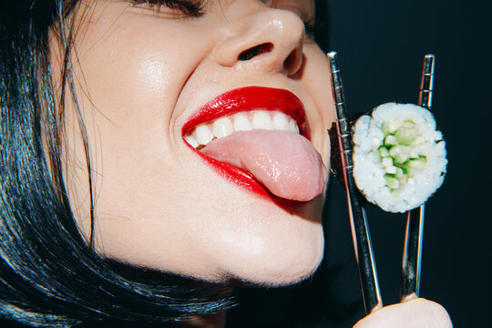 Playful woman teasingly tasting sushi with chopsticks, showcasing vibrant red lips and a confident expression against a dark background.
