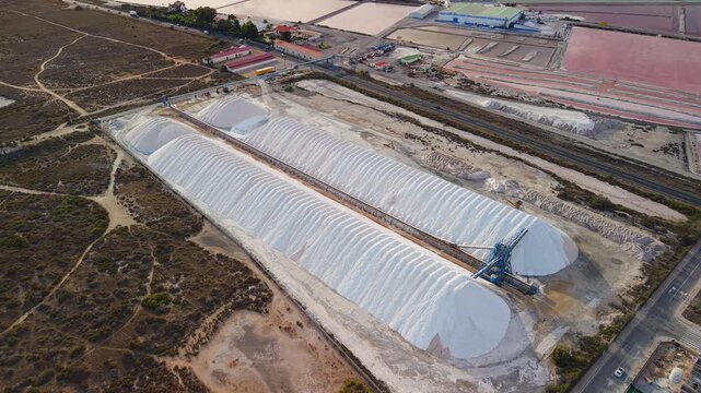 Aerial drone view of large salt piles at a production facility near Santa Pola, Spain, with colorful evaporation ponds and surrounding landscape.