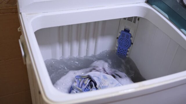 High-angle view inside a top-loading washing machine during the wash cycle. Clothes are swirling and agitating in soapy water with bubbles. Domestic household appliance in action.