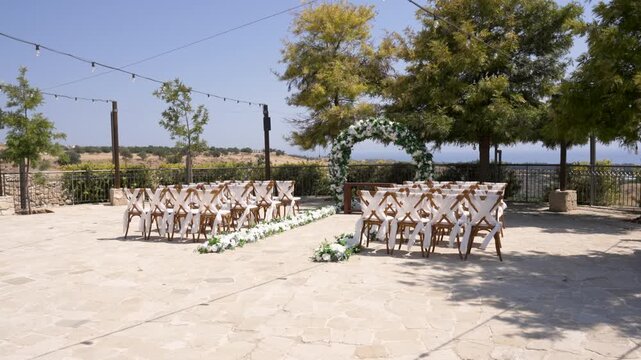 An elegant outdoor wedding setup features rows of chairs arranged for guests. The scene is adorned with floral decorations and set against a picturesque landscape under a clear sky.