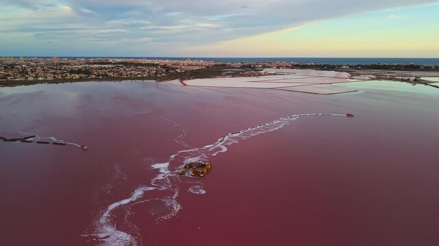 Aerial view of salt extraction barges operating on the pink waters of Laguna Salada de Torrevieja, Spain, with the city and salt mounds visible under colorful evening light.