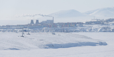 Fototapeta premium Winter Arctic landscape. View of the northern city. Anadyr, Chukotka, Russia. In the foreground are wind turbines and large fuel tanks at Cape Observation. Snowy tundra on the coast of a frozen river.