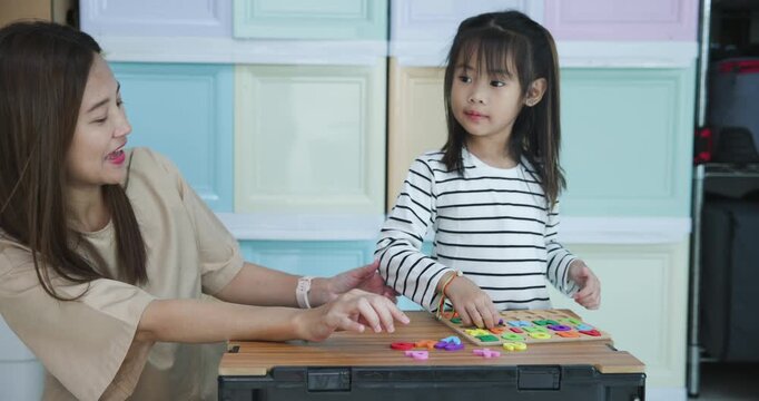  Mother and her young daughter are engaged in an educational activity at home, placing colorful letter and number pieces into a wooden puzzle board 