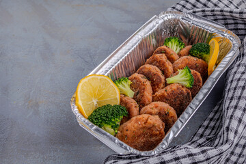 Salmon patties with lemon and broccoli in a foil baking container